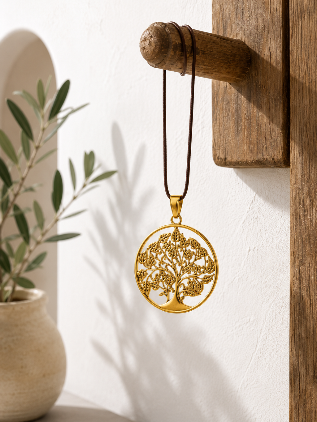 Colgante dorado Árbol de la Vida con cordón marrón, colgado sobre un soporte de madera en un interior luminoso.