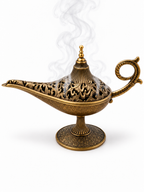 Lámpara mágica decorativa en acabado bronce envejecido con tapa calada y humo de incienso saliendo del interior sobre fondo blanco.